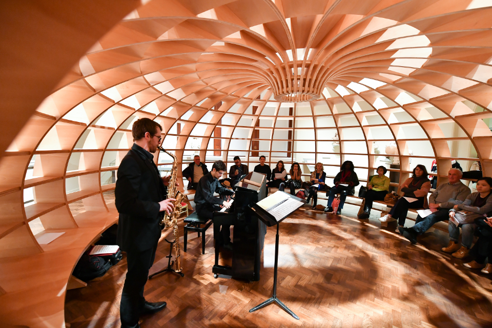 Two students performing the saxophone and the piano in a circular wooden room with light filling into the room, with the audience to the right, watching the students performing.